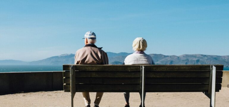 Zwei ältere Menschen sitzen auf einer Parkbank und sind von hinten zu sehen.