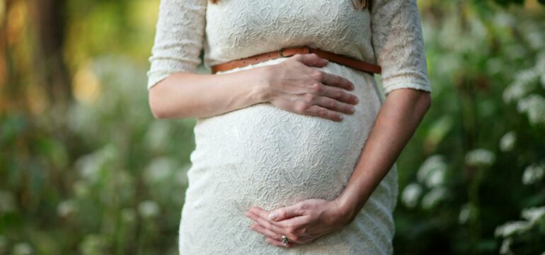 Eine schwangere Frau in einem weißen Kleid greift auf ihren Bauch.